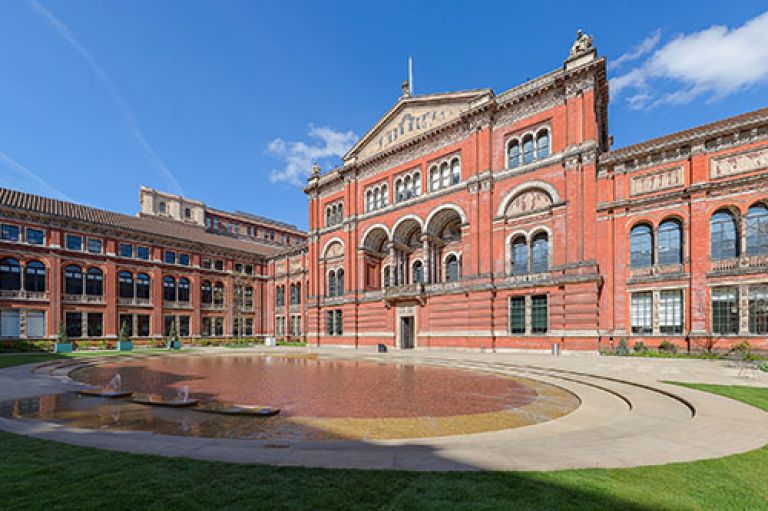 An image of the V&A Museum Central Garden. It is a red building with arches and Greek-resembling drawings of various figures below the arches. Towards the bottom of the image is a flat fountain. The sky is bright blue.