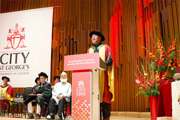 An image of Dr Gus Casely-Hayford OBE delivering a speech. He stands in the centre of the image at a red lectern with City St George's logo and strapline (The University of business, practice and the professions) and wears his graduating garb: a gown with yellow and red and a tam hat (a rounded floppy black hat for PhDs)/ To his right are academics sitting on stage in their own graduating garb. Behind them is a huge logo of City against the wooden pannelled wall of the Barbican, which looks like panels of wood and wood pipes in its design. To His right are large red and orange flowers.